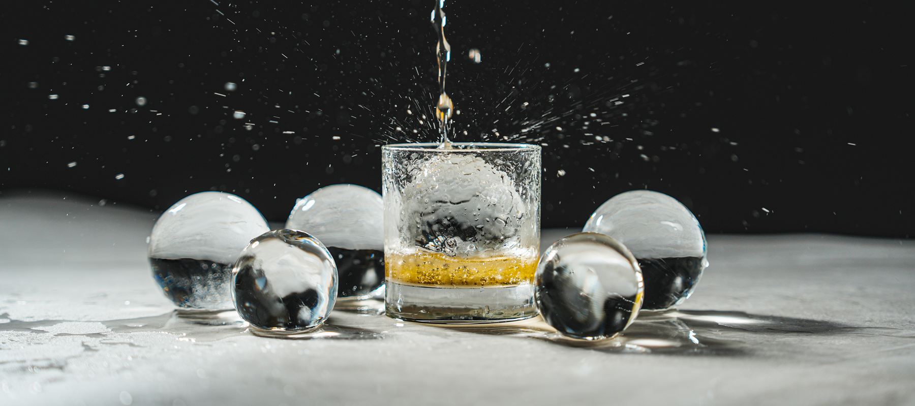 Clear ice spheres surrounding a glass with a clear ice spheres and a clear liquid is being poured in from a distance creating a splash.